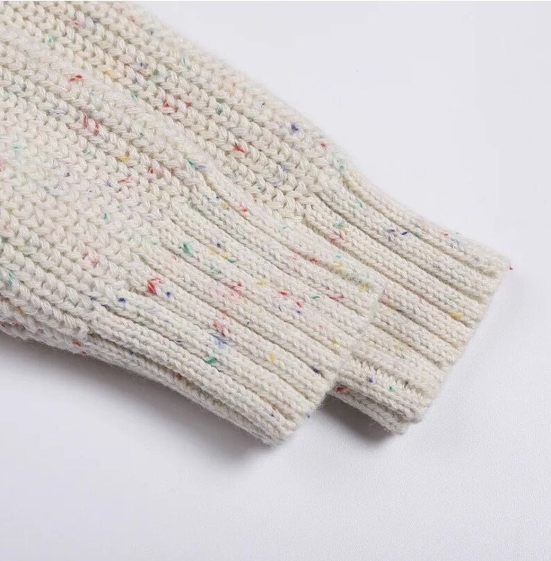 May include: Close-up of the cuff of a white knitted sweater with colorful flecks throughout the fabric.