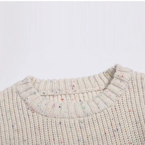 May include: Close-up of the neckline of a cream-colored sweater with a speckled pattern of multi-colored dots.