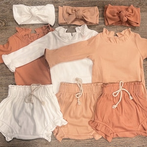 Könnte beinhalten: Drei Sets Babykleidung in Pfirsich, Weiß und Braun. Jedes Set enthält ein langärmliges Shirt mit Rüschenkragen und eine Rüschenhose. Die Hose hat einen Kordelzug. Es gibt auch drei Stirnbänder in Pfirsich, Weiß und Braun.