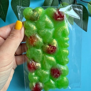May include: A green soap bar with a bubbly texture, infused with red and gold accents, is held in a clear plastic wrapper. The soap has a unique, organic shape and is set against a blue background.