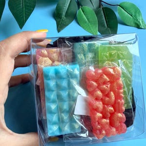 Può includere: Una scatola di plastica trasparente piena di saponi colorati. I saponi sono di varie forme e colori, tra cui rosa, blu, verde e rosso. I saponi hanno superfici strutturate e sono confezionati in cellophane trasparente.