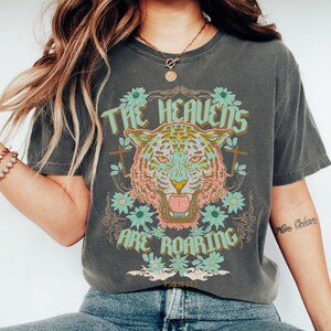 Puede incluir: Una camiseta gris con un gr&aacute;fico de un tigre con flores y el texto "The Heavens Are Roaring" y "Psalm 18:11".