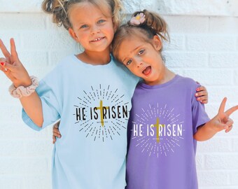 May include: Two children wearing light blue and purple t-shirts with the text "He is Risen" and a gold cross graphic. The children are smiling and making peace signs.