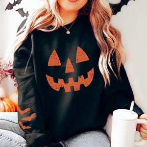 May include: Black Halloween sweatshirt featuring a jack-o'-lantern face in orange. The sweatshirt has orange bat graphics on the left sleeve. The image includes a white tumbler with a straw, pumpkins, and decorative bats.