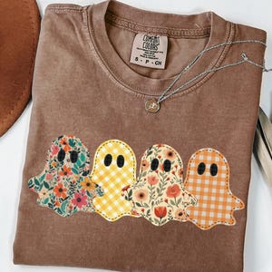 May include: Espresso-colored t-shirt featuring four ghost appliqués with floral and gingham patterns. The shirt has a crew neck and a Comfort Colors tag. A silver necklace with a small pendant is draped across the shirt.