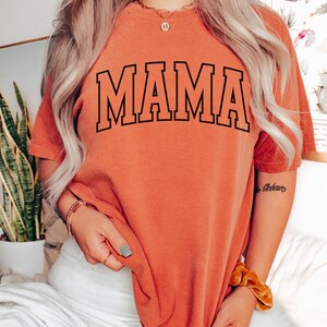 May include: An orange-red t-shirt with the word "MAMA" in large, black, block letters. The shirt is worn by a person with long, wavy blonde hair. The person is wearing white pants and gold jewelry.