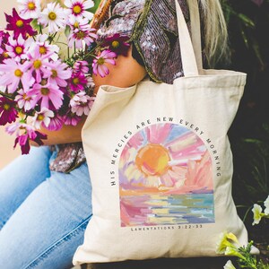 Pode incluir: Uma bolsa tote de lona branca com uma pintura de nascer do sol colorida e o texto "His mercies are new every morning" e "Lamentations 3:22-33".