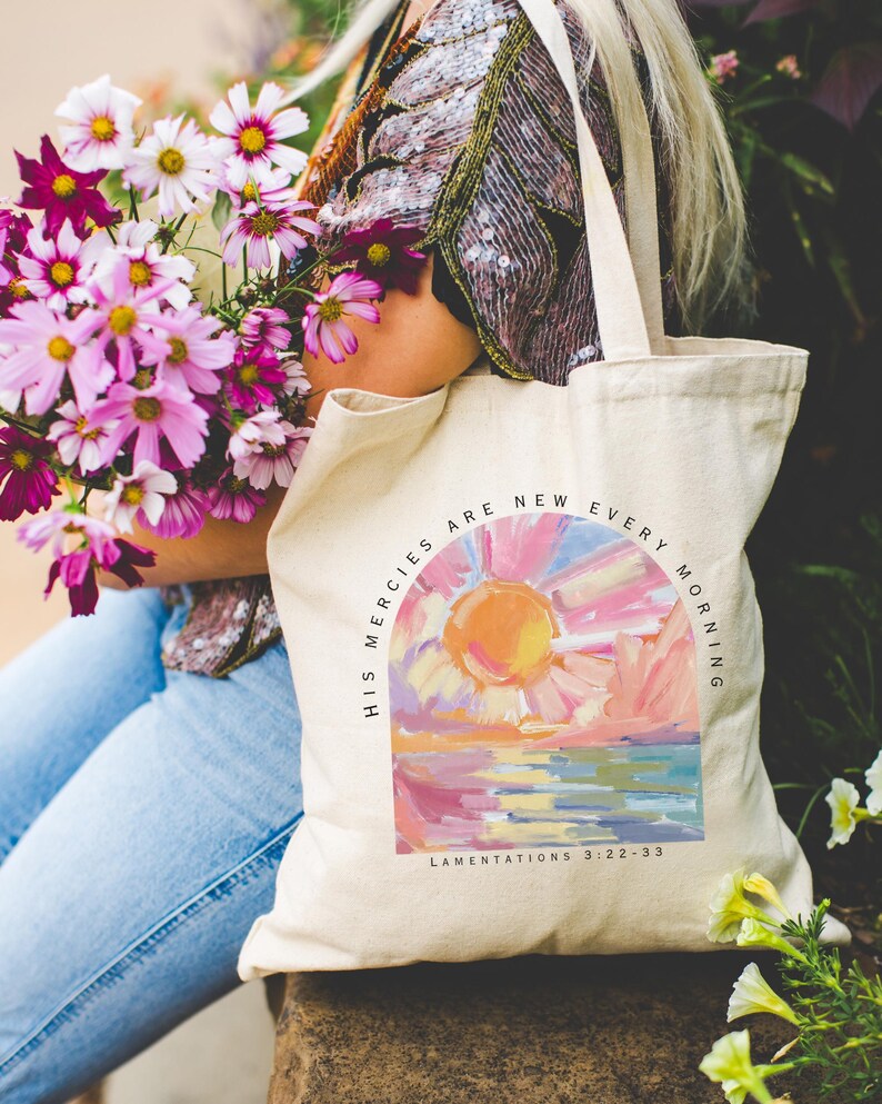 Pode incluir: Uma bolsa tote de lona branca com uma pintura de nascer do sol colorida e o texto "His mercies are new every morning" e "Lamentations 3:22-33".