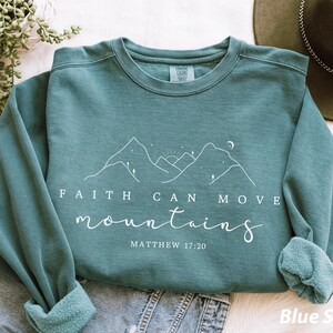 以下が含まれることがあります： 「Faith Can Move Mountains」の文字と山のグラフィックが描かれた、ティール色のスウェットシャツ。「Matthew 17:20」の文字が下にあります。スウェットシャツはジーンズの上に折りたたまれています。