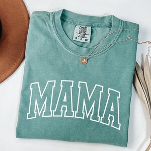 May include: A teal-colored t-shirt with the word "MAMA" in bold, white, block letters. A gold necklace with a pendant rests on the shirt. A brown hat and dried plant are also in the image.