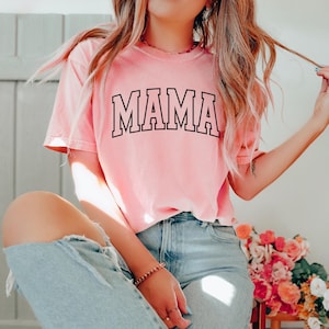 May include: A light pink t-shirt with the word "MAMA" in black block letters. The shirt is a crew neck style and appears to be made of a soft material. The shirt is paired with light blue jeans.