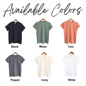 Puede incluir: Seis camisetas colgadas en perchas, mostrando diferentes colores: negro, verde musgo, coral, gris antracita, marfil y blanco. El texto "Available Colors" est&aacute; escrito encima de las camisetas.