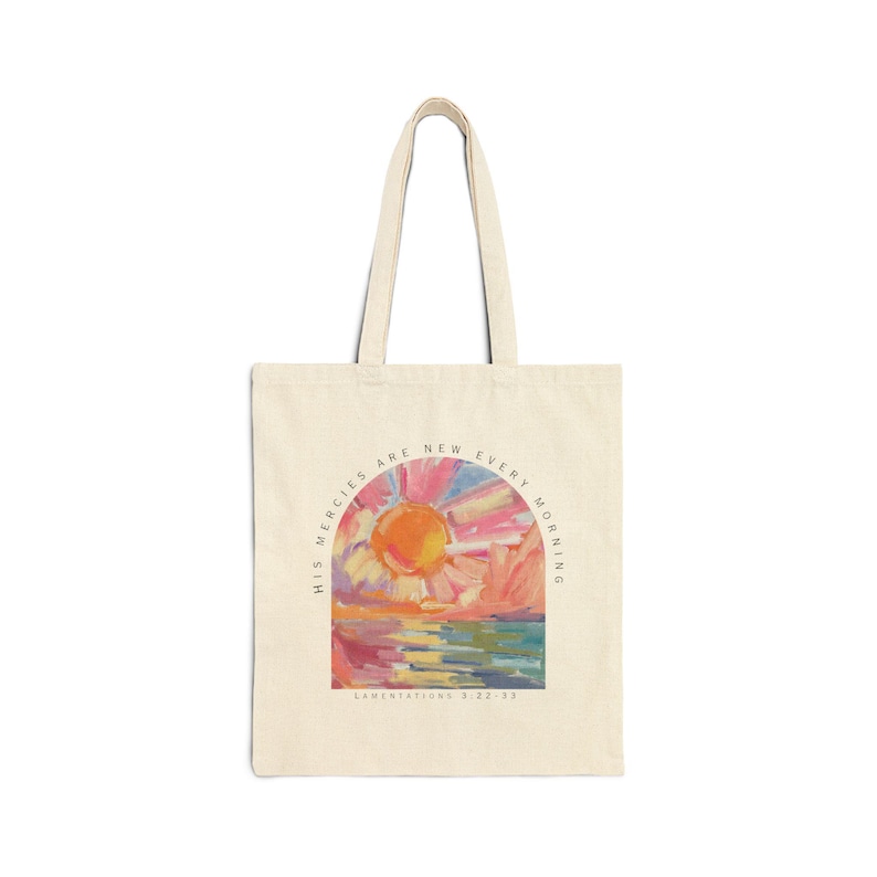Pode incluir: Uma bolsa de tecido bege com uma pintura de nascer do sol colorida e o texto "His mercies are new every morning" e "Lamentations 3:22-23".