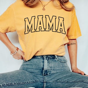 May include: Mustard yellow t-shirt with the word "MAMA" in bold, black, outlined letters. The shirt is worn with blue jeans and white sneakers. A gold necklace and bracelet are also visible.