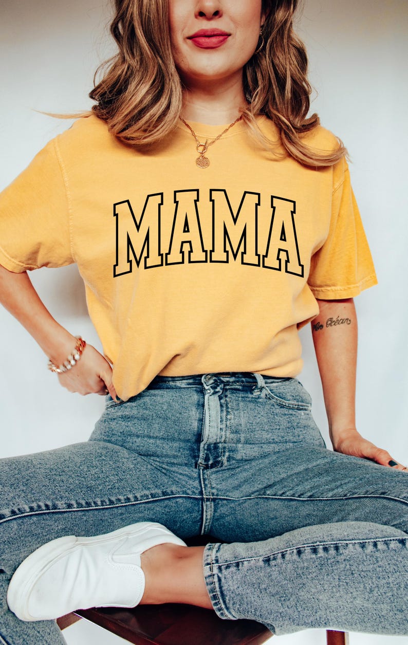 May include: Mustard yellow t-shirt with the word "MAMA" in bold, black, outlined letters. The shirt is worn with blue jeans and white sneakers. A gold necklace and bracelet are also visible.