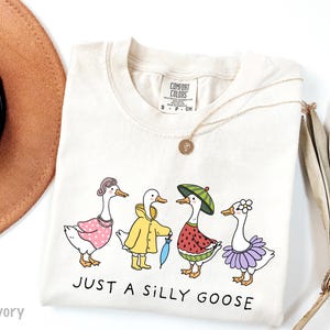 May include: A beige t-shirt with a graphic of four geese wearing different outfits. The geese are wearing a pink dress, a yellow raincoat, a watermelon costume, and a purple tutu. The text "JUST A SILLY GOOSE" is printed below the geese.