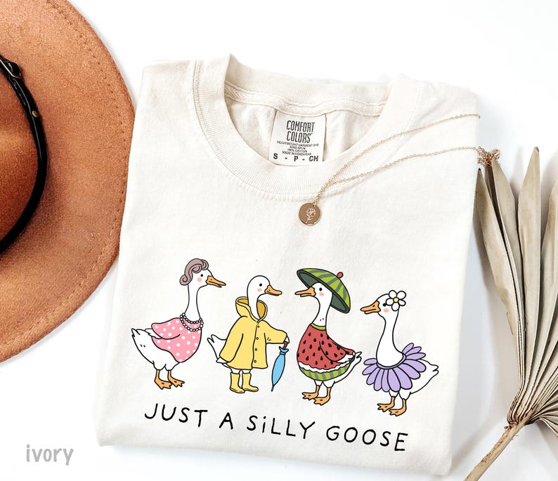 May include: A beige t-shirt with a graphic of four geese wearing different outfits. The geese are wearing a pink dress, a yellow raincoat, a watermelon costume, and a purple tutu. The text "JUST A SILLY GOOSE" is printed below the geese.