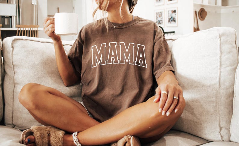 May include: A brown t-shirt with the word "MAMA" in white block letters. The shirt is worn by someone sitting on a light-colored couch, holding a white mug. The overall aesthetic is casual and relaxed.