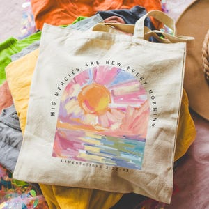 Pode incluir: Uma bolsa de lona branca com uma pintura de nascer do sol colorida e o texto "His mercies are new every morning" e "Lamentations 3:22-23".