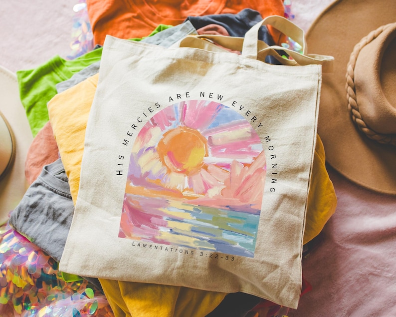 Pode incluir: Uma bolsa de lona branca com uma pintura de nascer do sol colorida e o texto "His mercies are new every morning" e "Lamentations 3:22-23".