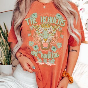 Puede incluir: Una camiseta de color &oacute;xido con un estampado gr&aacute;fico de un tigre rugiente con manchas verdes y amarillas. El gr&aacute;fico tambi&eacute;n incluye flores y el texto "THE HEAVENS ARE ROARING".