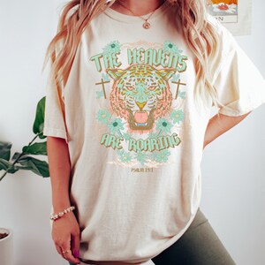 Puede incluir: Una camiseta beige con un gr&aacute;fico de un tigre con un dise&ntilde;o floral y el texto "The Heavens Are Roaring" y "Psalm 19:1".