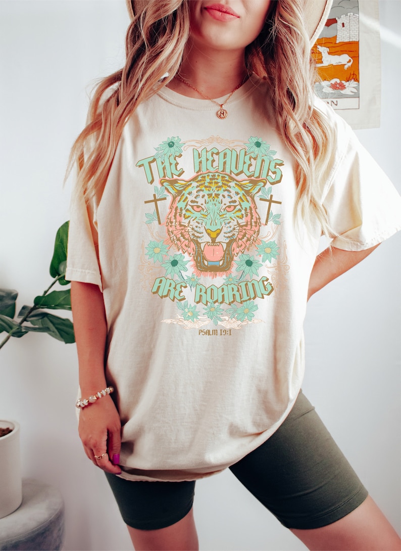 Puede incluir: Una camiseta beige con un gr&aacute;fico de un tigre con un dise&ntilde;o floral y el texto "The Heavens Are Roaring" y "Psalm 19:1".