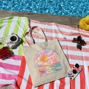 Pode incluir: Uma bolsa de lona branca com uma pintura colorida de um nascer do sol e o texto "His mercies are new every morning" e "Lamentations 3:22-23". A bolsa est&aacute; sobre uma toalha de praia listrada rosa e verde.