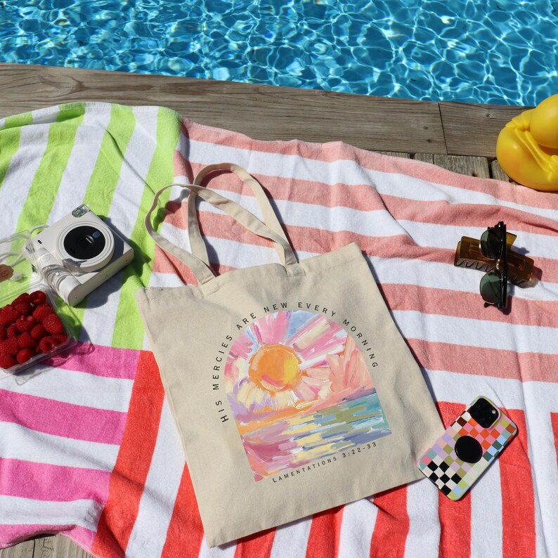 Pode incluir: Uma bolsa de lona branca com uma pintura colorida de um nascer do sol e o texto "His mercies are new every morning" e "Lamentations 3:22-23". A bolsa est&aacute; sobre uma toalha de praia listrada rosa e verde.