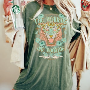 Puede incluir: Camiseta verde de gran tama&ntilde;o con un gr&aacute;fico de un tigre con el texto "The Heavens Are Roaring" y la escritura "Salmo 84:1".
