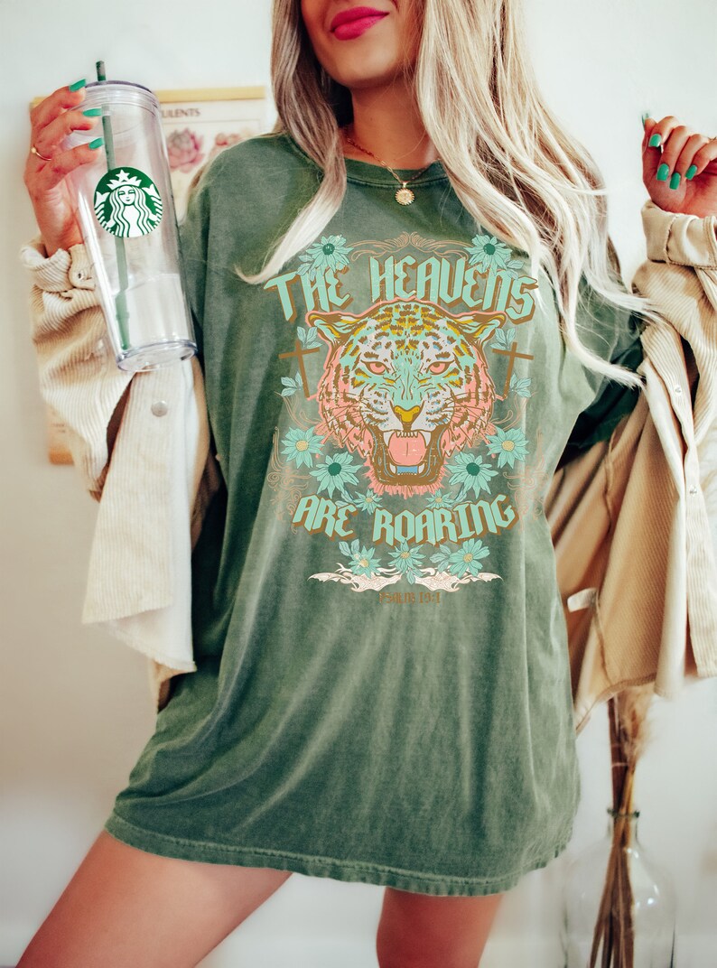 Puede incluir: Camiseta verde de gran tama&ntilde;o con un gr&aacute;fico de un tigre con el texto "The Heavens Are Roaring" y la escritura "Salmo 84:1".