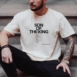 Peut inclure: T-shirt crème avec l'inscription noire "SON OF THE KING" et un verset biblique. La personne portant le t-shirt est assise sur des marches. Elle porte un pantalon noir et un bracelet.