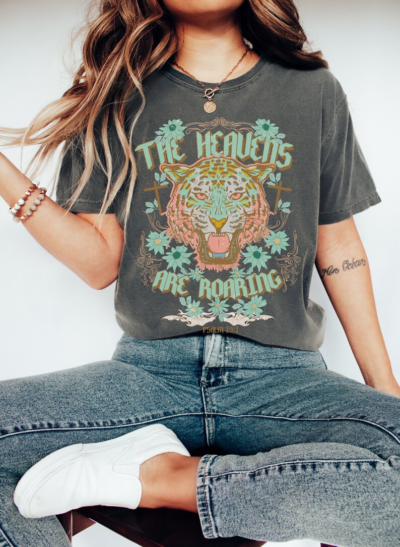 Puede incluir: Una camiseta gris con un gr&aacute;fico de un tigre con flores y el texto "The Heavens Are Roaring" y "Psalm 18:11".