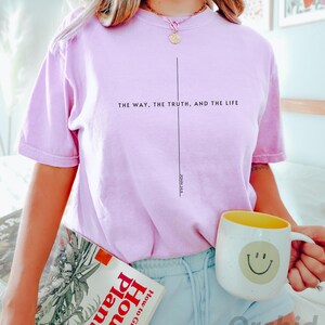 May include: A pink t-shirt with the text "THE WAY, THE TRUTH, AND THE LIFE" and "JOHN 14:6" printed on it. The shirt is being worn by a person holding a book and a mug with a smiley face.