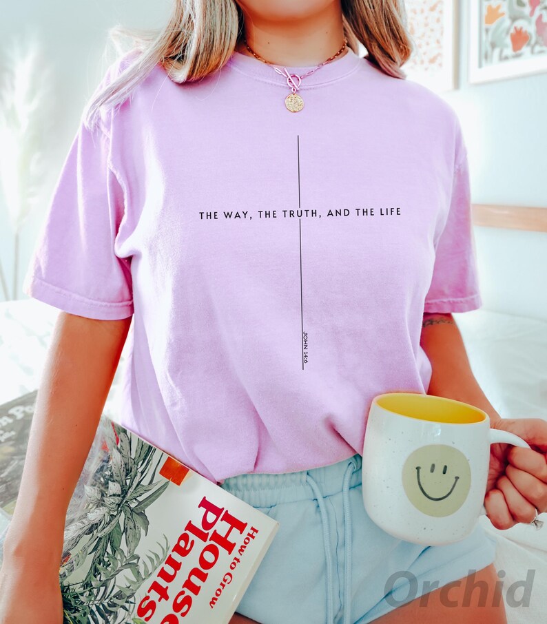 May include: A pink t-shirt with the text "THE WAY, THE TRUTH, AND THE LIFE" and "JOHN 14:6" printed on it. The shirt is being worn by a person holding a book and a mug with a smiley face.
