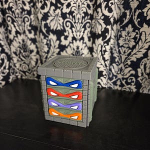 May include: A gray, square-shaped storage organizer designed to look like a sewer manhole structure. It features four stacked drawers, each with a different colored mask—blue, red, purple, and orange—covering the eyes of a character.