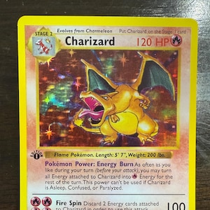 May include: A Charizard Pokémon card, Stage 2, with a holographic background. The card features a yellow border and a large illustration of a Charizard. The card text includes "120 HP", "Fire Spin", and "Energy Burn".