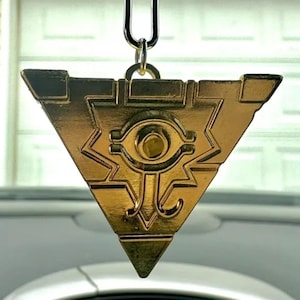 May include: A gold-colored triangular pendant with an eye symbol hanging from a silver chain. The pendant is hanging from a rearview mirror in a car.