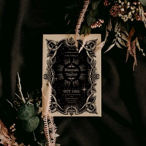 Puede incluir: Una invitaci&oacute;n de boda de estilo vintage con elementos de dise&ntilde;o celestiales en blanco y negro. La invitaci&oacute;n presenta los nombres "Hannah y Taylor" y la fecha "OCT 13th". La invitaci&oacute;n est&aacute; rodeada de flores secas y vegetaci&oacute;n.