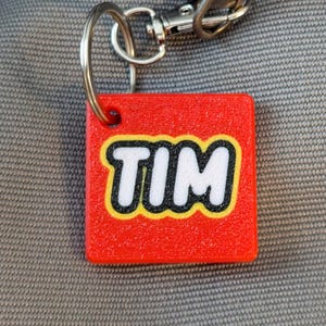 May include: Red plastic keychain with a white name "TIM" in a black outline. The keychain is attached to a silver metal ring.