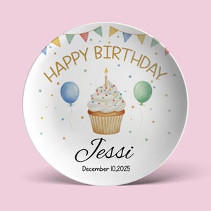 May include: White ceramic plate with a birthday theme. The plate features a cupcake with a lit candle, balloons, and the text "HAPPY BIRTHDAY" in gold. It also includes the name "Jessi" and the date "December 10, 2025". Colorful confetti dots and a banner complete the design.
