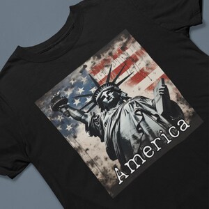 May include: Black t-shirt featuring a graphic of the Statue of Liberty with an American flag background. The word "America" is printed in white across the bottom of the image. The design is distressed, giving it a vintage look.