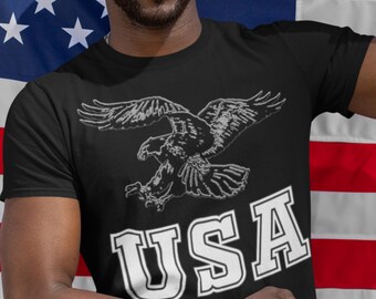 Puede incluir: Camiseta negra con un diseño de águila blanca en vuelo y la palabra "USA" en letras blancas y gruesas. El diseño está centrado en la camiseta. El águila es detallada.
