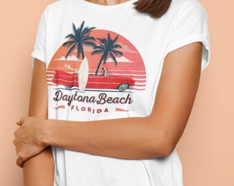 Puede incluir: Camiseta blanca con un diseño retro que presenta un descapotable rojo, una tabla de surf y palmeras sobre un fondo de puesta de sol. El texto "Daytona Beach FLORIDA" se muestra en una fuente vintage.