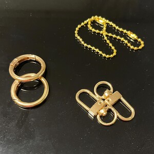 以下が含まれることがあります： ゴールドカラーの金属製金具。2つの丸い留め金、チェーン、Dリングが2つ付いた留め金が含まれています。暗い表面に配置されており、光沢のある反射面が特徴です。バッグやジュエリー用のアクセサリーです。