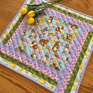 May include: A quilted table runner with a light blue background featuring a pattern of bunnies, eggs, and flowers. The runner has a multi-coloured border with stripes of green, pink, and purple.