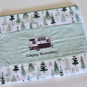 May include: A green and white zippered pouch with a camping trailer design and the text "Camping Reservations". The pouch has a white background with a green watercolor pattern of pine trees.