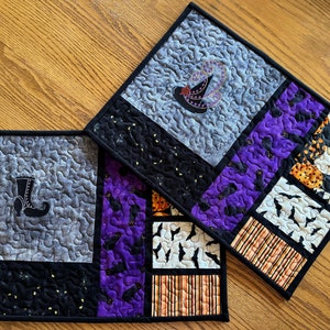 May include: Two quilted placemats with a Halloween theme. One placemat features a witch's hat and the other features a witch's boot. Both placemats are made of different fabrics with a patchwork design.
