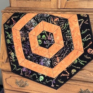 Peut inclure: Chemin de table sur le thème d'Halloween avec un motif géométrique. Le chemin de table présente un hexagone orange central entouré de tissu noir avec un motif répété de crânes et de squelettes. Les bords sont également noirs avec la même impression effrayante.