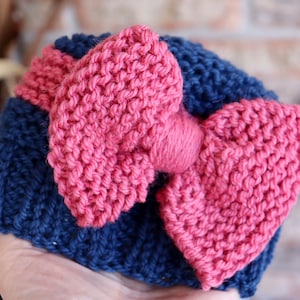 Peut inclure: Un bandeau en tricot avec un grand nœud rose. Le bandeau est bleu marine et le nœud est d'une nuance de rose plus foncée.